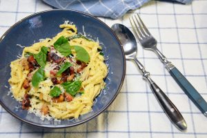 Cremige Pasta mit Speck, Spinat und geriebenem Parmesan in einer blauen Schale auf einem blau karierten Geschirrtuch