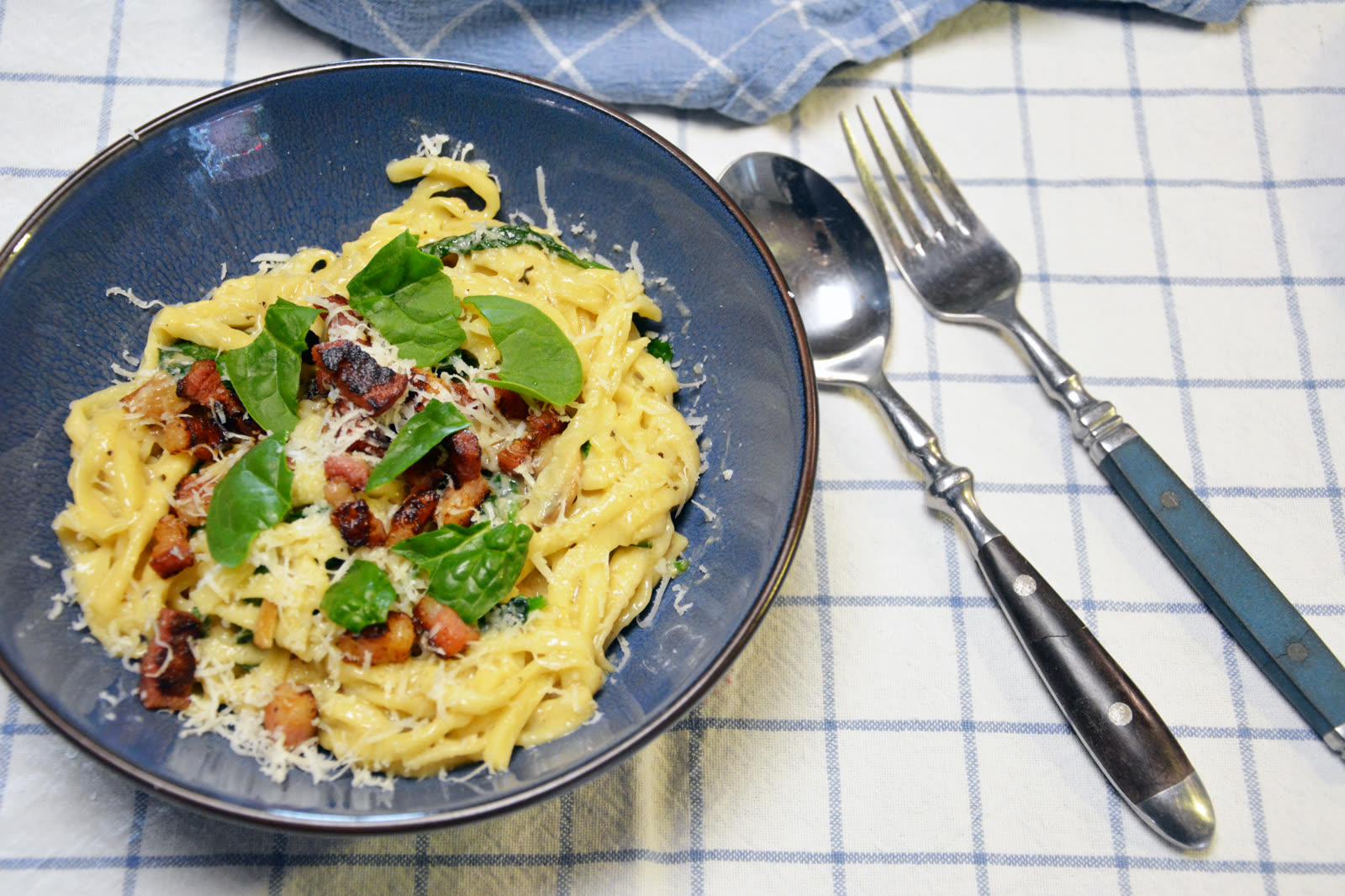 Cremige Pasta mit Speck, Spinat und geriebenem Parmesan in einer blauen Schale auf einem blau karierten Geschirrtuch