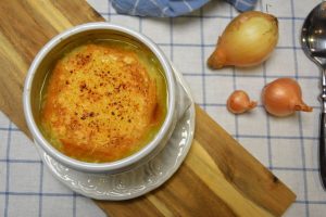 Französische Zwiebelsuppe in einer weißen Suppentasse mit überbackener Käsebrotscheibe, auf einem Holzbrett neben ganzen Zwiebeln