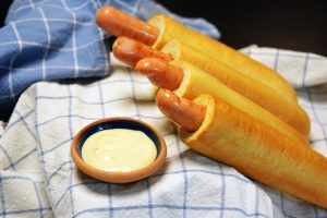 Vier fertige French Hotdogs mit der Wurst im Loch liegen auf einem blau karierten Geschirrtuch neben einer kleinen Schale mit heller Dressing