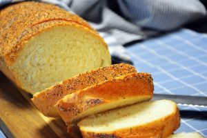 Selbst gebackenes Weißbrot mit Mohn auf der Kruste, in Scheiben geschnitten auf einem Holzbrett neben einem blau karierten Geschirrtuch