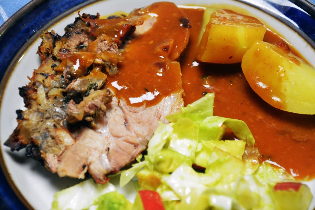 Zwei Scheiben gefüllter Schweinenacken mit dunkler Pilzfüllung zwischen den Scheiben auf einem Teller mit blauem Rand, dazu braune Soße, Salzkartoffeln und grüner Salat