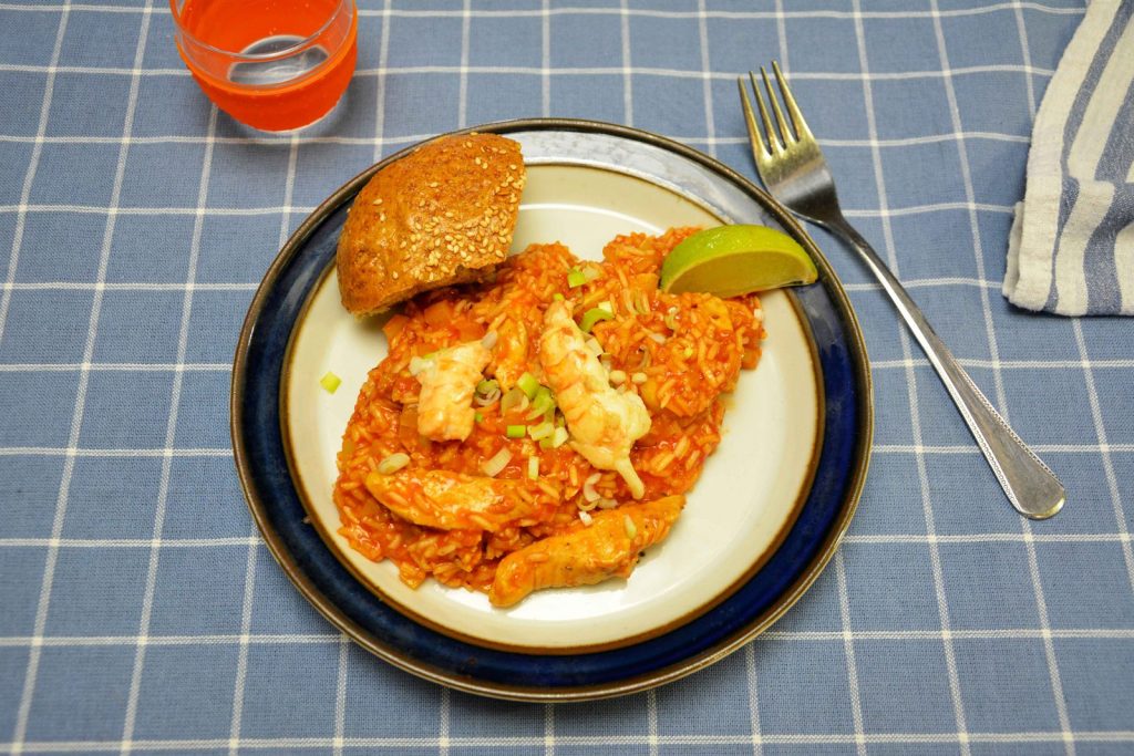 Jambalaya mit rotem Tomatenreis, Hähnchenstreifen und Kaisergranat-Schwänzen auf einem weißen Teller mit Limettenspalte, gehackter Frühlingszwiebel und einem Sesambrötchen