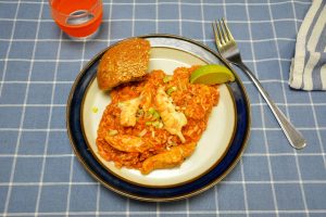 Jambalaya mit rotem Tomatenreis, Hähnchenstreifen und Kaisergranat-Schwänzen auf einem weißen Teller mit Limettenspalte, gehackter Frühlingszwiebel und einem Sesambrötchen