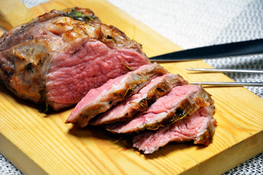 Ein gebräunter Braten auf einem hellen Holzbrett, teilweise in rosa Scheiben aufgeschnitten, mit ganzen Kräuterzweigen auf der Kruste, daneben ein Tranchiermesser mit schwarzem Griff und eine Gabel