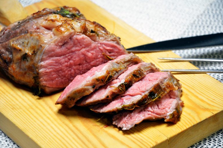 Ein gebräunter Braten auf einem hellen Holzbrett, teilweise in rosa Scheiben aufgeschnitten, mit ganzen Kräuterzweigen auf der Kruste, daneben ein Tranchiermesser mit schwarzem Griff und eine Gabel