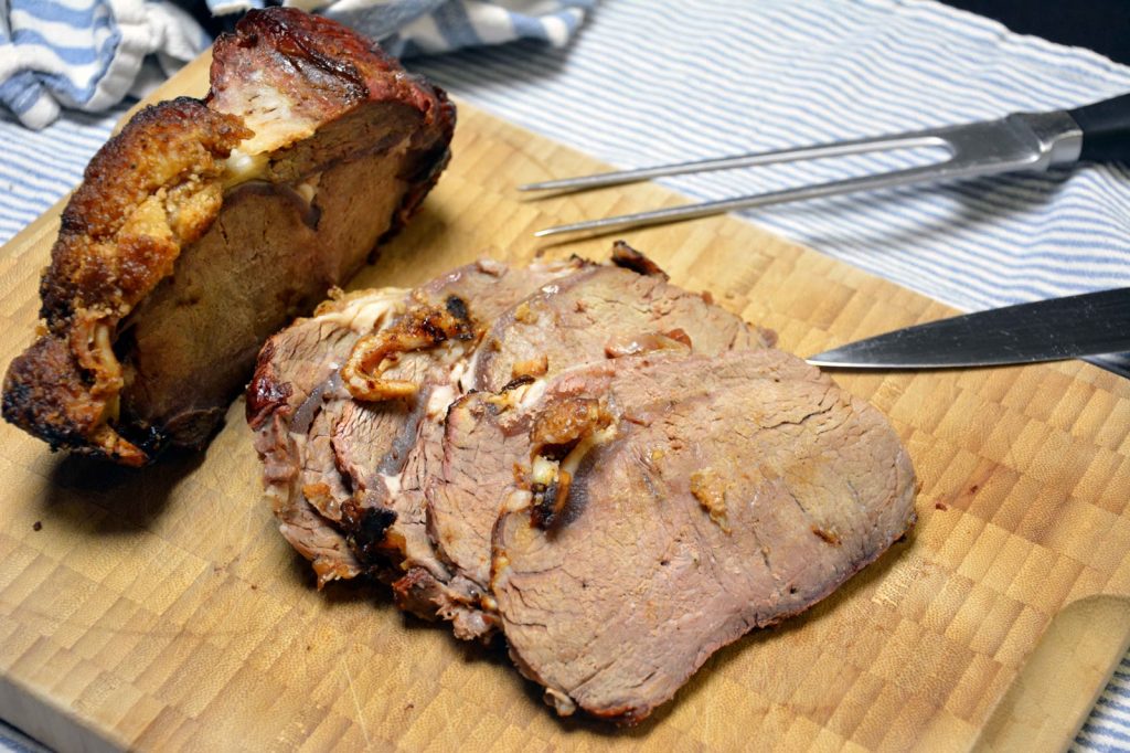 Ein im Ganzen gegrillter Braten auf einem hellen Schneidebrett aus Hirnholz, mit knuspriger, goldbrauner, rautenförmig eingeschnittener Fettkruste obenauf, zur Hälfte in dicke Scheiben geschnitten mit heller, leicht rosa-grauer Mitte, daneben eine Tranchiergabel und ein Tranchiermesser, im Hintergrund ein blau-weiß gestreiftes Geschirrtuch
