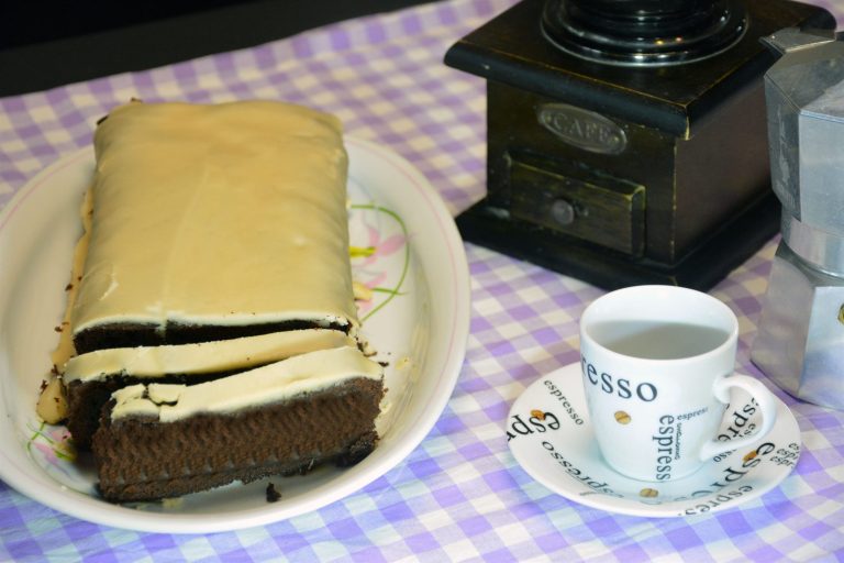 In Scheiben geschnittener Kaffeekuchen mit heller Kaffeeglasur auf einem weißen Teller, daneben eine Espressotasse, eine alte Kaffeemühle und eine Mokkakanne