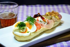 Ein halbes aufgeschnittenes Baguette liegt offen auf einem langen, rechteckigen weißen Servierteller; vorn Eierstücke mit einem Klecks Mayonnaise und Kresse, dann Tomatenscheiben mit einem Dillzweig, dann eine Scheibe Hamburgerryg mit Erbsen-Möhren-Salat und einem Stück Aspik, dahinter Leberpastete und Salami mit Röstzwiebeln und eine Scheibe saure Gurke; links ein Glas Bier, darunter eine lila-weiß karierte Tischdecke vor dunklem Hintergrund