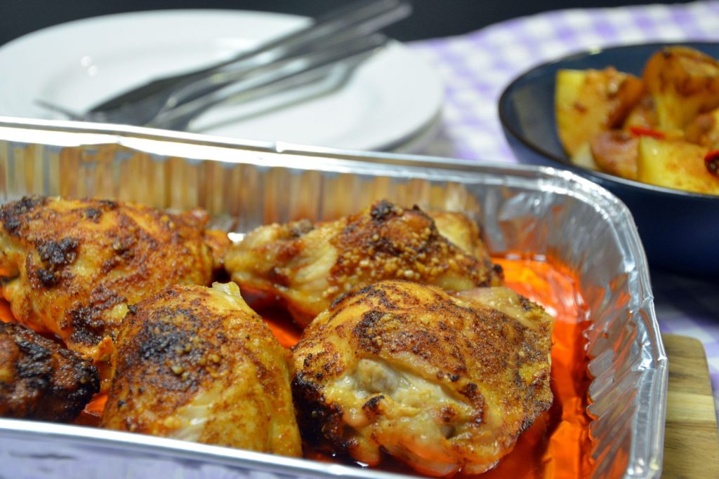 Vier goldbraune, mit Paprika gewürzte Hähnchen-Oberkeulen mit Haut in einer rechteckigen Aluminiumschale, unten eine rotorange Marinade; dahinter ein weißer Teller mit Messer und Gabel, oben rechts eine dunkelblaue Schüssel mit gebackenen Kartoffelstücken und einem Stück roter Chili, auf einer lila-weiß karierten Tischdecke vor dunklem Hintergrund