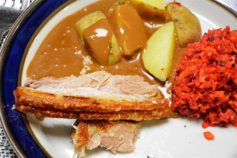 Zwei Scheiben langzeitgegarter Krustenbraten mit goldener, knuspriger, geschichteter Schwarte und hellem Fleisch auf einem weißen Teller mit dunkelblauem Rand; dahinter halbierte und geviertelte Kartoffeln mit Schale unter hellbrauner Soße, rechts ein Häufchen fein gehackter Rohkostsalat in Rot und Orange