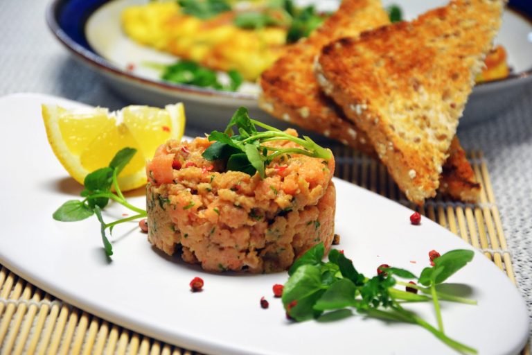 Ein rundes Türmchen Lachstatar mit Dill und rosa Pfefferbeeren, gekrönt von Brunnenkresse, auf einem länglichen weißen Teller mit einer Zitronenspalte und lose verstreuten rosa Pfefferbeeren; dahinter Dreiecke aus geröstetem Kernbrot und ein Teller mit Omelett; darunter eine Bambusmatte