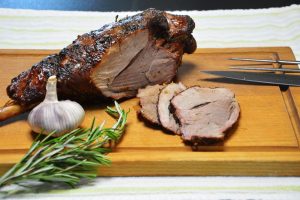 Eine gegrillte Lammkeule mit Knochen, dessen Ende links herausragt, auf einem Holzschneidebrett über einem gestreiften Leinentuch; dunkle, fast schwarze Rauchkruste, ein Schnitt im Fleisch zeigt das rosa Innere, drei Scheiben liegen vorn rechts; links im Vordergrund eine ganze Knoblauchknolle und Rosmarinzweige, rechts ein Messer und eine Vorlegegabel