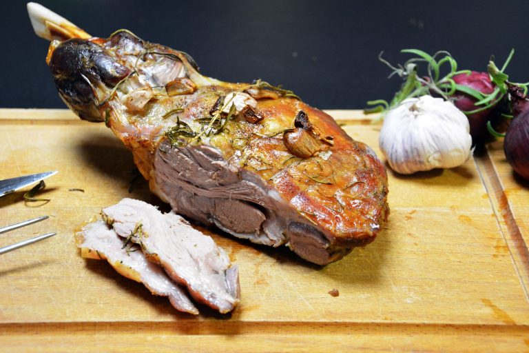 Eine im Ofen gebratene Lammschulter mit Knochen, der links herausragt, auf einem Holzschneidebrett; knusprig braune Oberfläche mit gerösteten Knoblauchzehen und Rosmarinnadeln; zwei Scheiben rosa Fleisch liegen angeschnitten davor; links ein Messer und eine Gabel, rechts eine ganze Knoblauchknolle, rote Zwiebeln und Rosmarinzweige vor dunklem Hintergrund