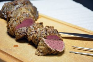 Ein bei Niedrigtemperatur gegartes Rinderfilet, in dicke Scheiben geschnitten, auf einem hellen Holzbrett; das Fleisch ist gleichmäßig rosa bis an die braune Kruste. Rechts liegen ein Tranchiermesser und eine Tranchiergabel mit zwei langen Zinken, dahinter ein weißes Geschirrtuch, oben rechts ist der Hintergrund dunkel